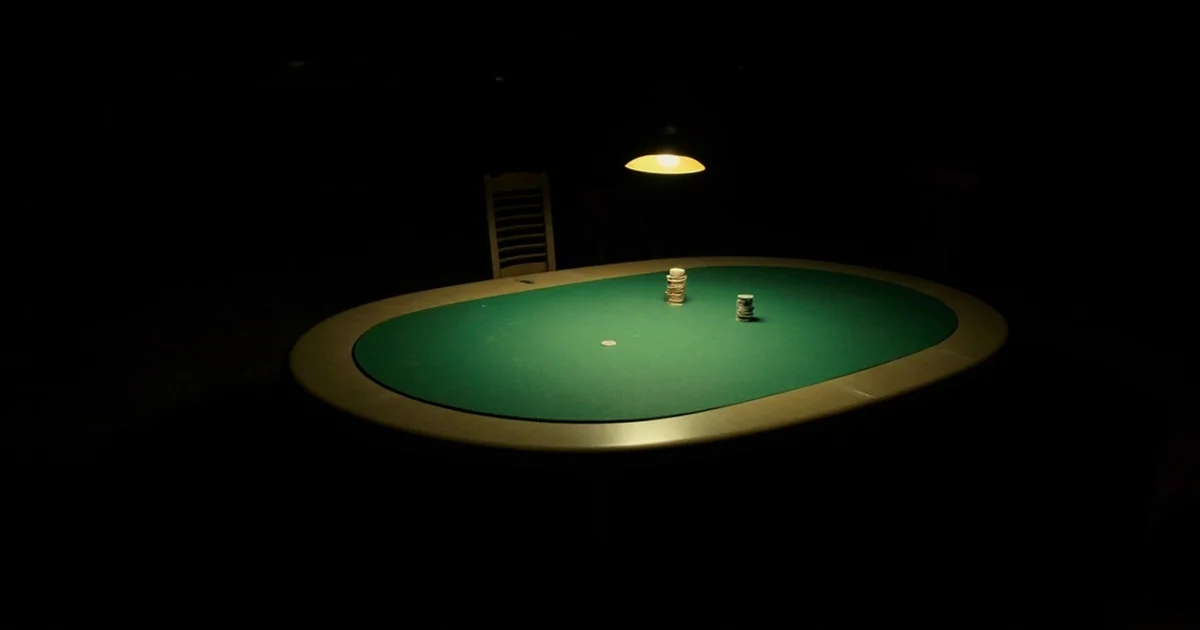 Empty green-felt poker table with a pushed-back chair, stacked chips, and an amber overhead light.