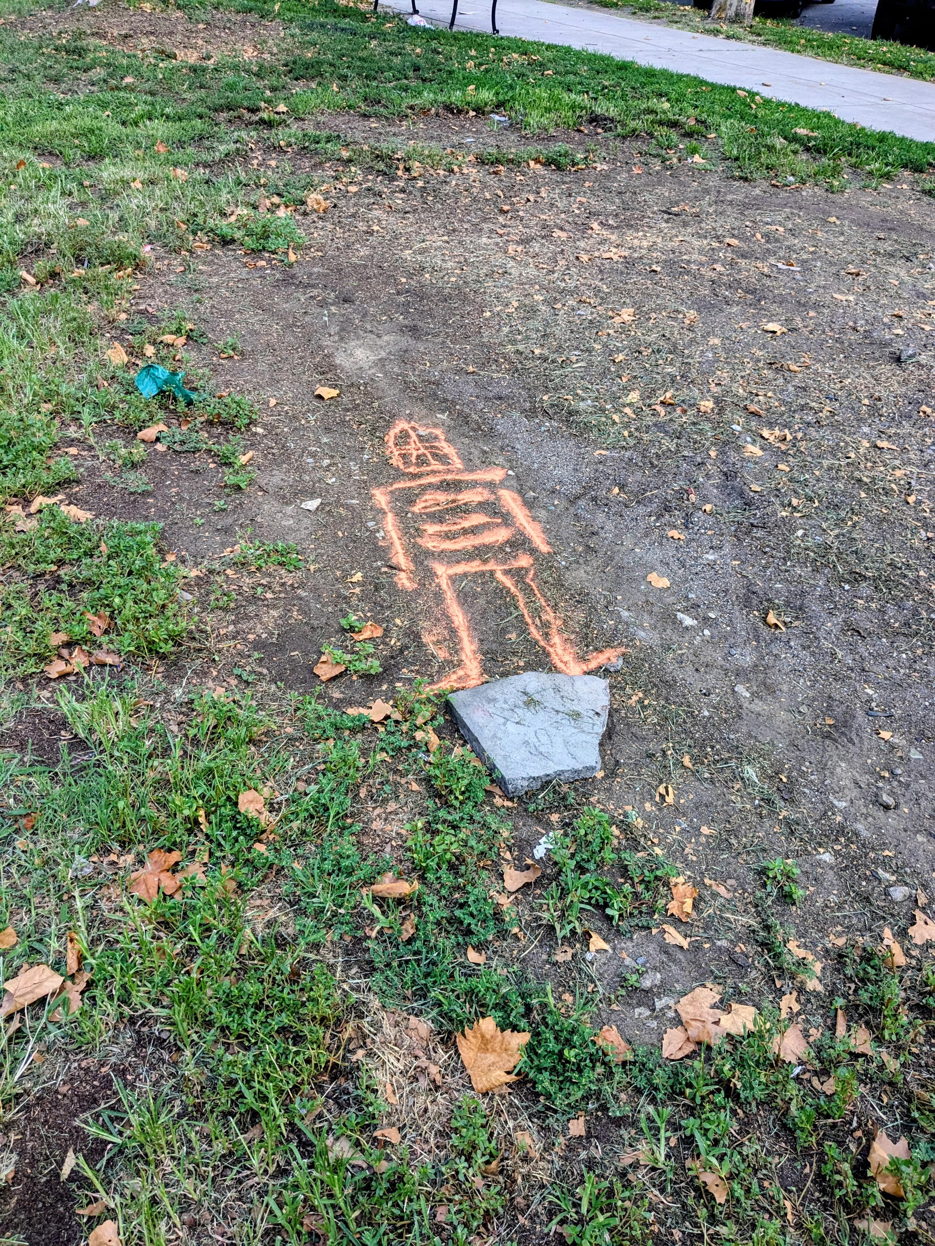 An orange robot-like stick figure drawn into a patch of bare dirt beside a sidewalk in the Fairmount neighborhood of Philadelphia
