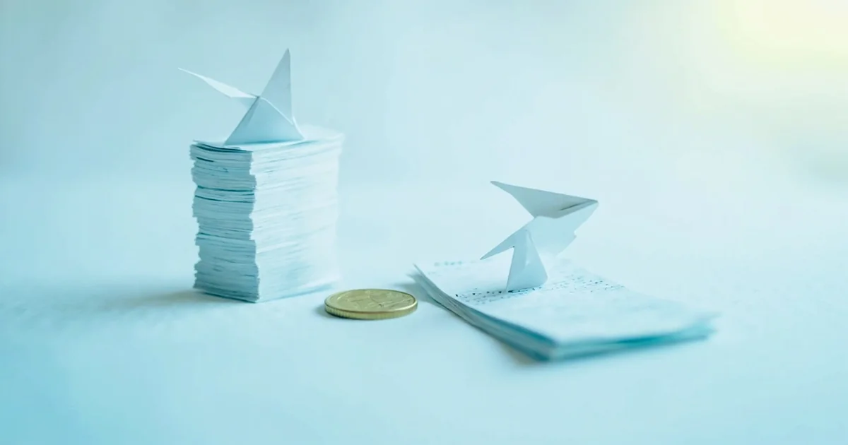 Uneven stacks of pay stubs with small paper airplanes balanced on top, separated by a single coin on a white surface.